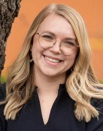 Eine blonde Frau mit Brille und schwarzem Oberteil lächelt und lehnt sich gegen einen Baumstamm vor orangefarbenem Hintergrund.