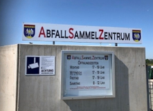 Ein Schild für Abfall Sammel Zentrum mit Öffnungszeiten auf einer Betonwand. Das Schild ist weiß mit roten Buchstaben und hat ein Logo. Unter dem Schild befindet sich ein kleineres weißes Schild mit Öffnungszeiten in Deutsch.