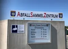Ein Schild für Abfall Sammel Zentrum mit Öffnungszeiten auf einer Betonwand. Das Schild ist weiß mit roten Buchstaben und hat ein Logo. Unter dem Schild befindet sich ein kleineres weißes Schild mit Öffnungszeiten in Deutsch.