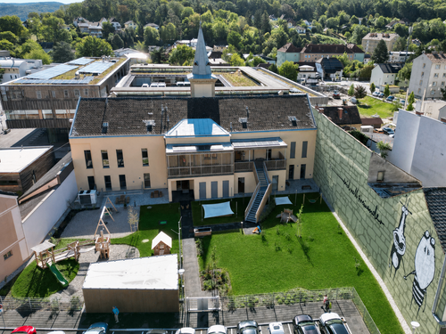 Luftaufnahme eines modernen Gebäudekomplexes mit einem Turm, umgeben von einem grünen Rasen und einem Spielplatz. Das Gebäude hat eine Mischung aus beige und blauen Dächern.