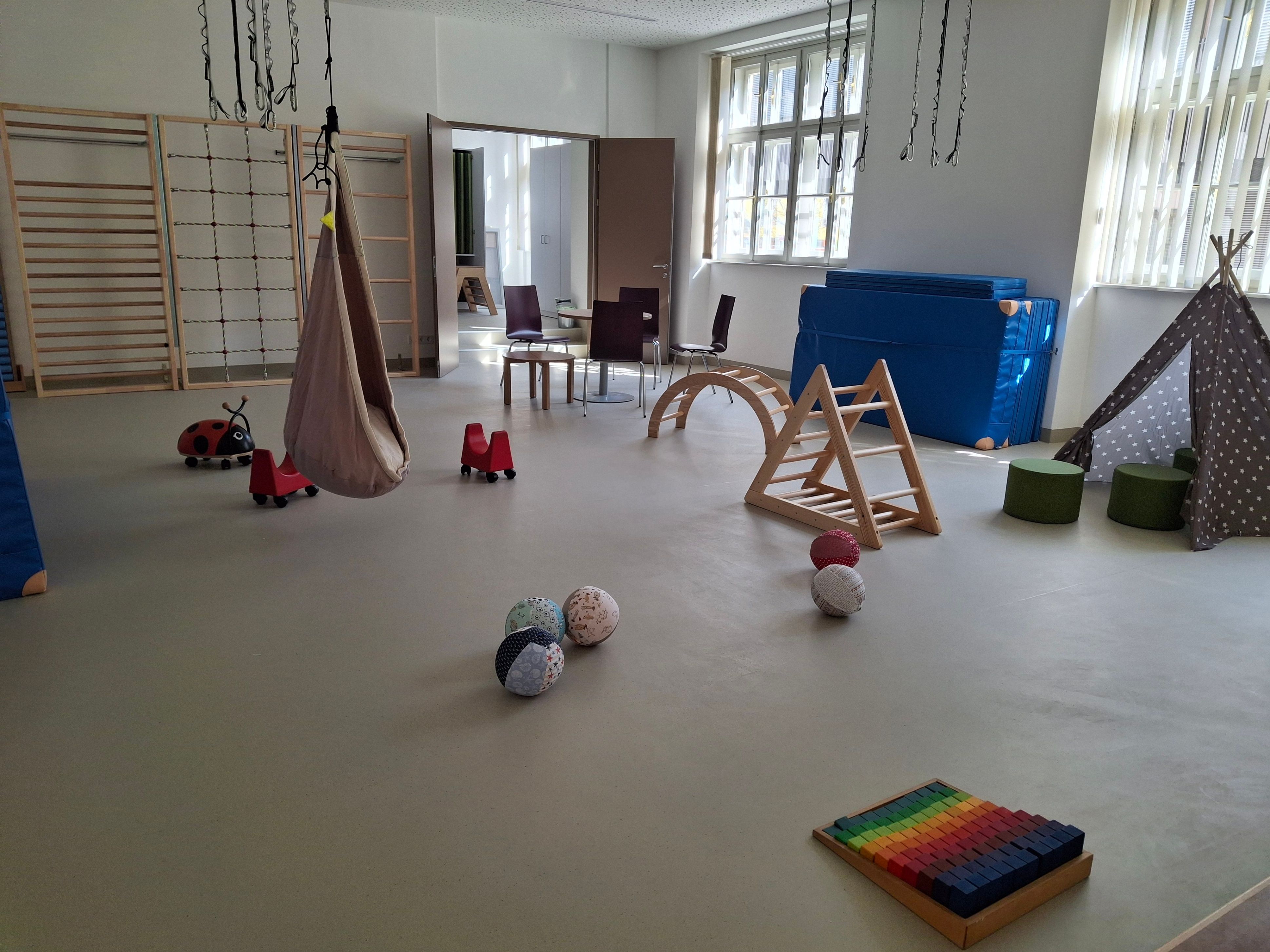 Ein Spielzimmer mit verschiedenen Spielzeugen und Spielgeräten. Ein Schaukel hängt an der Decke und ein Regenbogen-Xylophon liegt auf dem Boden.