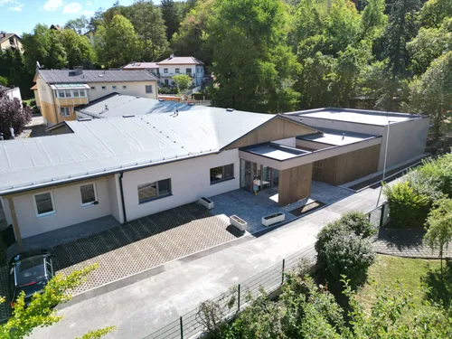 Luftaufnahme eines modernen Gebäudekomplexes mit grauem Dach und Einfahrt, umgeben von Bäumen und anderen Gebäuden.