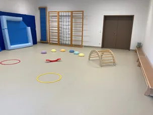 Ein Spielzimmer mit weißem Boden, verschiedenen farbigen Spielzeugen, einer hölzernen Kletterleiter und einem blauen aufblasbaren Objekt.