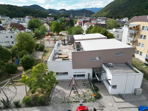 Ein Luftbild eines modernen Gebäudekomplexes mit einem Spielplatz davor, umgeben von Bäumen und Häusern, mit Bergen im Hintergrund.