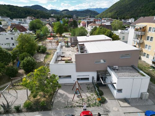 Ein Luftbild eines modernen Gebäudekomplexes mit einem Spielplatz davor, umgeben von Bäumen und Häusern, mit Bergen im Hintergrund.