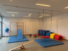 Ein Indoor-Spielbereich mit verschiedenen Spielzeugen und Ausrüstungen. Eine blaue Matte liegt auf dem Boden, ein blauer Ball steht auf einem Ständer und eine Rutsche befindet sich links. Eine weiße Tür befindet sich in der Mitte und einige Spielzeuge sind rechts.