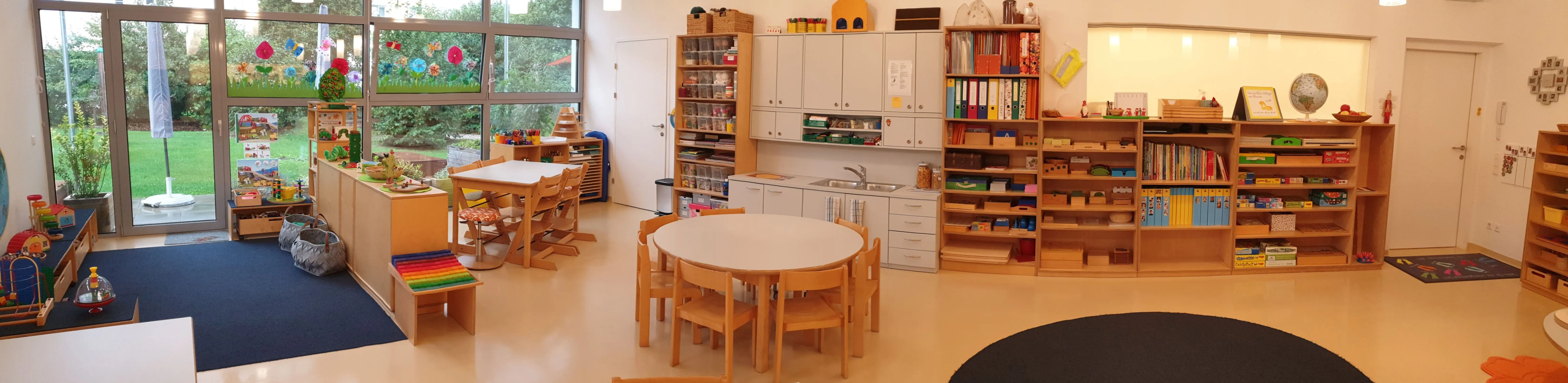 Ein Innenraum einer gut beleuchteten Raum, möglicherweise ein Klassenzimmer, mit Holztischen und Stühlen um einen zentralen Tisch herum angeordnet. Der Raum hat große Glasfenster mit bunten Zeichnungen, Holzregalen gefüllt mit Büchern und Spielzeug, und ein Waschbecken mit einem Wasserhahn.