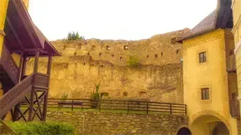 Eine alte Steinburg mit gelber Wand und einer Holztreppe. Es gibt einen Steinzaun mit einer Bank, einem Topf und einem Holzgeländer. Bäume und Berge sind in der Ferne.