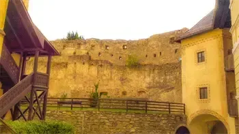 Eine alte Steinburg mit gelber Wand und einer Holztreppe. Es gibt einen Steinzaun mit einer Bank, einem Topf und einem Holzgeländer. Bäume und Berge sind in der Ferne.