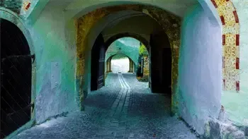 Ein schmaler Kopfsteinpflasterweg führt durch eine Reihe von gewölbten Durchgängen mit grünen und blauen Wänden. Die Bögen werden von Holztüren flankiert. Der Weg ist leer und zeigt ein helles Licht am Ende.