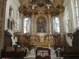 Ein kunstvoller Altar in einer Kirche mit einem zentralen Gemälde, flankiert von Statuen und umgeben von kunstvollen Holzarbeiten. Der Altar ist mit Blumen und einem Kreuz geschmückt.