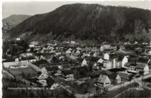 Ein Schwarz-Weiß-Foto zeigt eine kleine Stadt in einem Tal, mit Hügeln und Bergen im Hintergrund. Die Stadt besteht aus einer Mischung von Wohngebäuden, einige mit sichtbaren Schornsteinen, und einigen größeren Strukturen. Das Bild ist mit '1920' datiert.