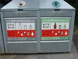 Ein Recyclingbehälter mit zwei Fächern. Jedes Fach hat ein Schild mit einer Warnung, es sauber zu halten, zusammen mit Symbolen für verschiedene recycelbare Gegenstände. Das obere Fach hat ein blaues Schild mit dem Wort 'Weissglas', während das untere Fach ein grünes Schild mit dem Wort 'Blauglas' hat.