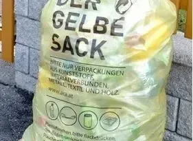 Ein gelber Plastiksack mit der Aufschrift 'Der Gelbe Sack' für recycelbare Materialien wie Kunststoffe, Metalle, Textilien und Holz. Der Sack hat eine Website-Adresse darauf.