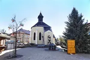 Eine kleine Kirche mit schwarzem Dach und Glockenturm befindet sich auf einem Stadtplatz, mit einem Baum und einer gelben Struktur auf der rechten Seite.