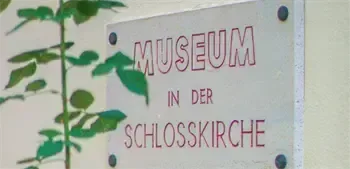 Ein weißes Schild mit rotem Text an einer Wand steht 'Museum in der Schlosskirche'. Ein Pflanzen mit grünen Blättern ist auf der linken Seite des Schildes sichtbar.