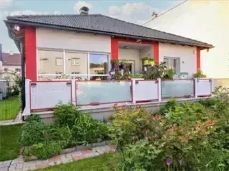 Ein weiß-rotes Haus mit schwarzem Dach und Glasfenstern. Das Haus hat einen Balkon mit Glasgeländern. Es ist von einem üppigen Garten mit grünem Rasen und verschiedenen Pflanzen umgeben.