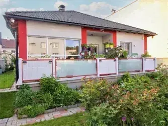Ein weiß-rotes Haus mit schwarzem Dach und Glasfenstern. Das Haus hat einen Balkon mit Glasgeländern. Es ist von einem üppigen Garten mit grünem Rasen und verschiedenen Pflanzen umgeben.