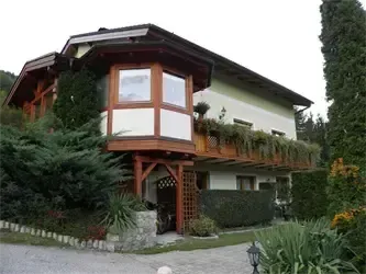Ein zweistöckiges Haus mit einem Balkon voller Pflanzen und Grün, umgeben von verschiedenen Büschen und Blumen, in einer malerischen Gegend gelegen.