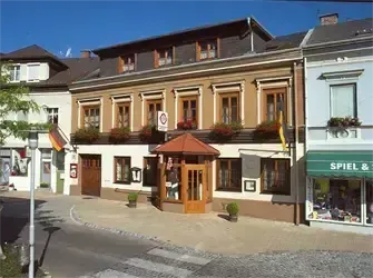 Ein Blick auf ein Gebäude mit vielen Fenstern und einer Holztür. Das Gebäude hat ein grünes Vordach und ein Schild mit der Aufschrift 'SPIEL & '. Draußen stehen Pflanzen und eine Fahne.