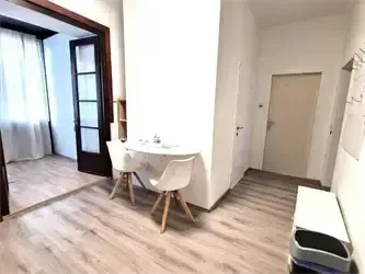 Ein leerer Raum mit Holzboden, weißen Wänden und einem kleinen Tisch mit zwei Stühlen. Es gibt eine weiße Tür, ein Fenster mit Vorhängen und einen Mülleimer neben der Tür.