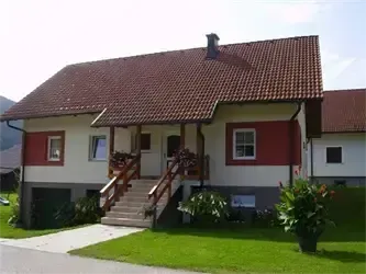 Ein zweistöckiges Haus mit rotem Dach und weißen Wänden, eine Treppe führt zu einer Tür, und davor ein Garten mit grünem Rasen und Pflanzen.
