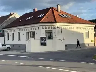 Eine Person geht an einem Gebäude mit dem Schriftzug 'Adria Pizzeria & Restaurant' vorbei. Das Gebäude hat ein rotes Dach und weiße Wände. Ein weißes Auto ist davor geparkt.