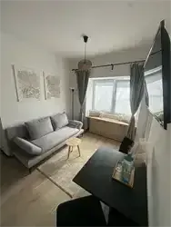 Ein minimalistisches Wohnzimmer mit einem grauen Sofa, einem Couchtisch und einem Fenster mit grauen Vorhängen. Der Raum ist mit einem Holzboden und weißen Wänden ausgestattet.