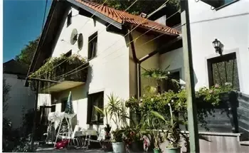 Ein weißes Haus mit braunem Dach und Balkon hat vorn Topfpflanzen und eine Lampe an der Wand. Ein Gartenschirm steht draußen.
