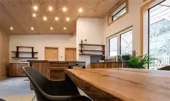 Eine moderne Küche mit Holzschränken, einem großen Holztisch und schwarzen Stühlen. Der Raum hat weiße Wände, eine Holzdecke und große Fenster.