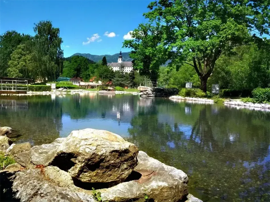 Ein ruhiger See mit großen Felsen und Bäumen spiegelt den blauen Himmel und ein entferntes Schloss an einem sonnigen Tag wider.