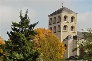 Eine Kirche mit einem hohen Turm und einem Kreuz darauf, umgeben von Bäumen und Laub mit Herbstfarben.