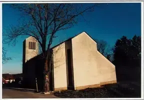 Ein Foto einer kleinen Kirche mit einem Turm und einer Person davor. Das Gebäude ist weiß mit einem Turm auf der linken Seite. Bäume umgeben die Kirche, und der Himmel ist klar.