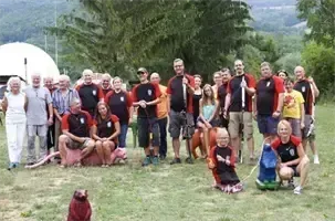 Eine Gruppe von Menschen in einheitlichen Kleidungsstücken, einige mit Hüten, posiert für ein Foto auf einer Rasenfläche. Im Hintergrund ist eine weiße Kuppel zu sehen. Bäume und ein Berg sind in der Ferne sichtbar.