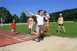 Ein Mädchen in einem weißen T-Shirt springt in eine Sandgrube, während mehrere Kinder auf einer Laufbahn zuschauen. Im Hintergrund sind Bäume und ein Torpfosten zu sehen.