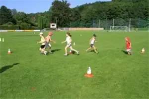 Eine Gruppe von Kindern läuft eine Staffel auf einem grünen Feld mit orangefarbenen Hütchen und einem Fußballtor im Hintergrund.