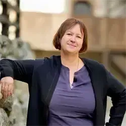Eine Frau mit kurzen Haaren und einem lilafarbenen Shirt steht gegen eine Steinmauer und lächelt. Sie trägt eine schwarze Jacke und eine Halskette. Dahinter ist ein verschwommener Hintergrund eines Gebäudes.