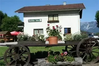 Ein weißes Haus mit braunem Dach und Blumen davor. Ein Wagen mit einem braunen Topf und Pflanzen steht auf dem Rasen. Berge und Bäume sind im Hintergrund.