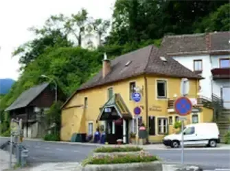 Ein gelbes Haus mit braunem Dach steht an einer Straßenecke. In der Nähe ist ein weißer Lieferwagen geparkt. Grüne Bäume und andere Gebäude sind im Hintergrund.
