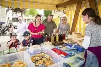 Eine Familie steht unter einem gelben Zelt mit einem Tisch voller Essen. Eine erwachsene Frau in einer lila Schürze serviert der Familie Essen. Auf dem Tisch befinden sich Kuchen, Brot und Flaschen mit Sirup.