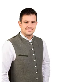 Ein Mann trägt eine graue Weste mit weißen Knöpfen und einem weißen Hemd. Er lächelt und scheint für ein Foto zu posieren.