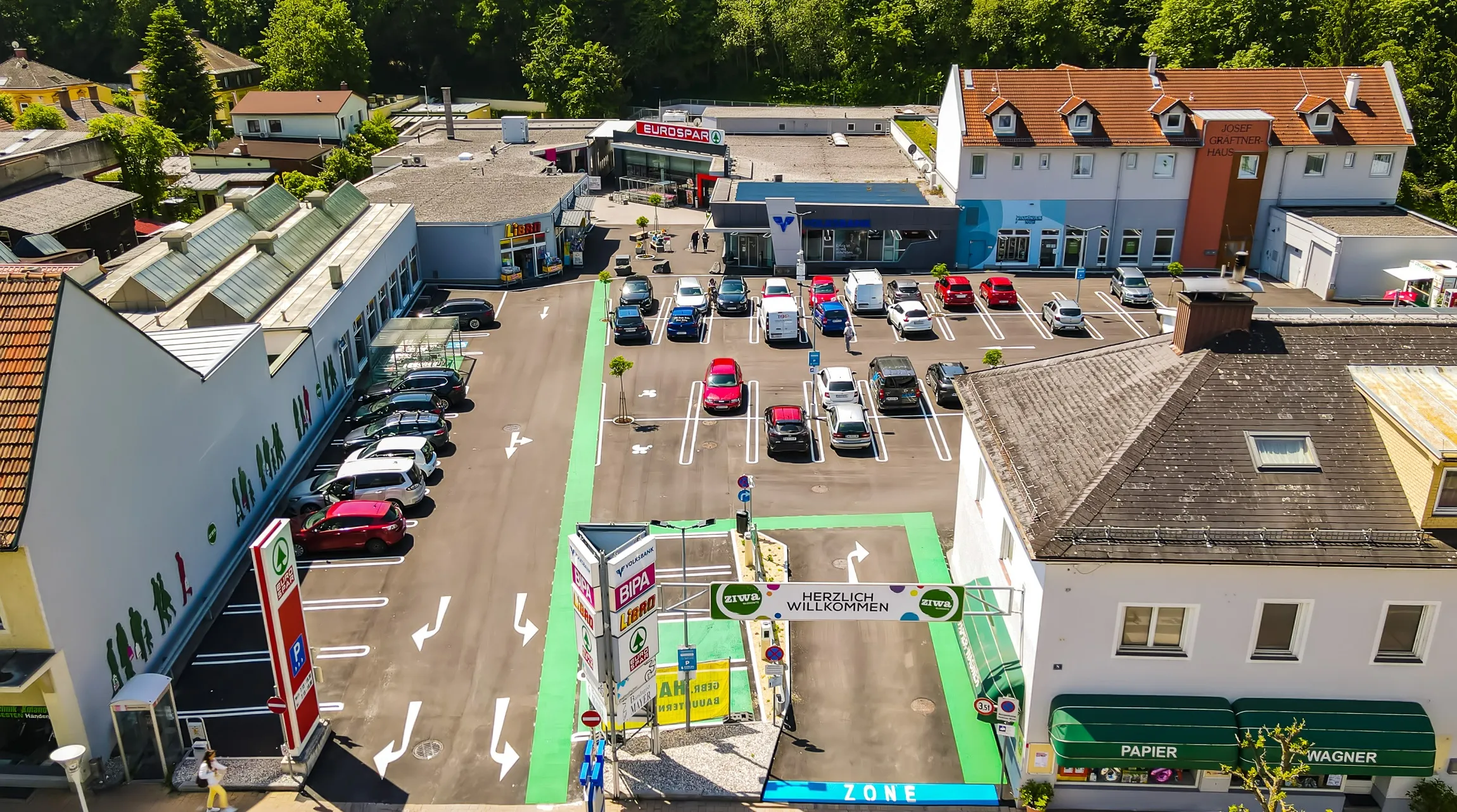 Luftaufnahme eines geschäftigen Parkplatzes mit mehreren Autos in Reihen, einem EuroSpar-Geschäft und einem Gebäude mit einem blauen Schild. Grüne und weiße Linien markieren die Parkplätze, und ein Schild steht Herzlich Willkommen.