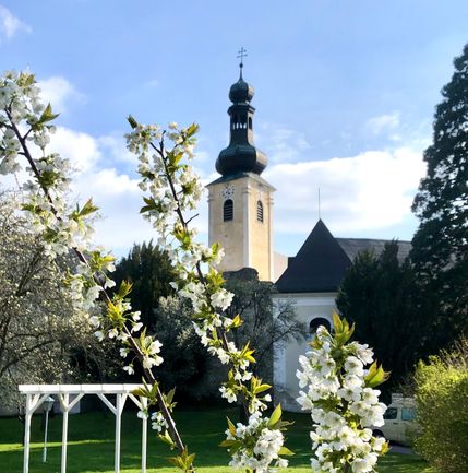 Eine Kirche mit Turm und Dachreiter ist von Bäumen und Gras umgeben. Die Kirche hat ein schwarzes Dach und eine weiße Wand.