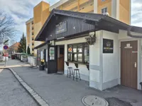 Die Vorderseite einer Bar namens Zum Wurstelbaron mit zwei Hockern draußen. Sie befindet sich an einer Straße mit einem Gebäude im Hintergrund.