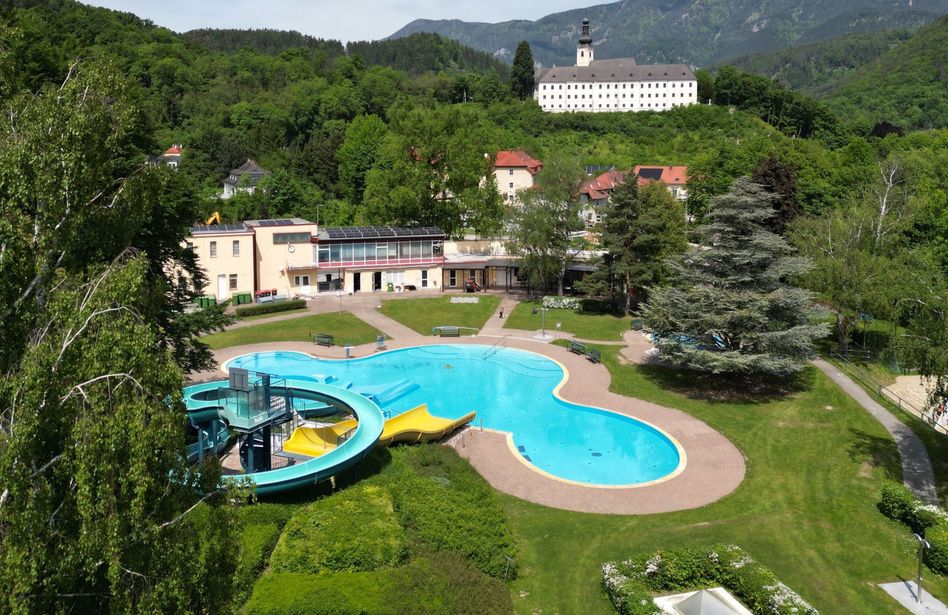 Eine Luftaufnahme eines großen Außenschwimmbeckens mit einer Wasserrutsche und einem Gebäude mit Solarmodulen auf dem Dach.