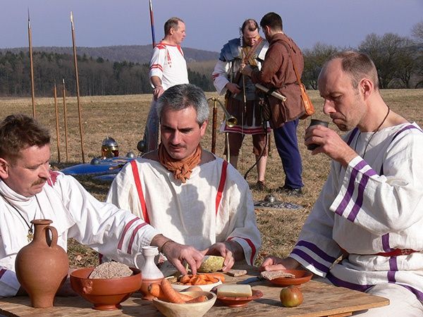 Vier Männer in antiker römischer Kleidung sitzen an einem Tisch und essen. Dahinter stehen zwei Männer in Rüstung mit Speeren.