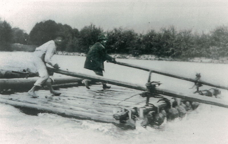Zwei Männer überqueren einen Fluss auf einer selbstgebauten Holzfloß. Sie halten sich an einem langen Stab fest.