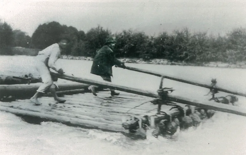 Zwei Männer überqueren einen Fluss auf einer selbstgebauten Holzfloß. Sie halten sich an einem langen Stab fest.