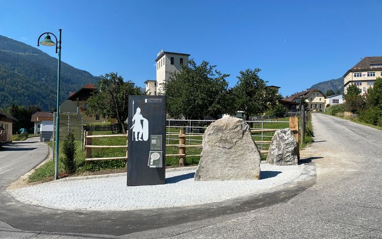 Ein schwarzes Informationsschild steht neben einem großen Felsen, mit einer Silhouette eines Wikingers mit Speer. Die Umgebung umfasst einen Holzzaun, eine Straße und Gebäude im Hintergrund.