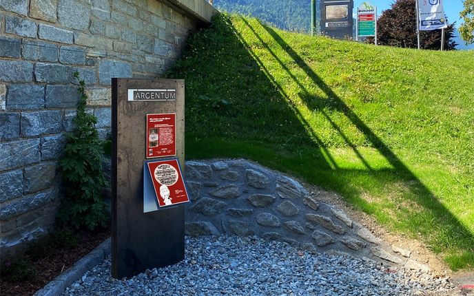 Ein Schild mit dem Wort Argentum ist an einer Steinmauer montiert. Es hat ein rotes Plakat mit einem Comic-Charakter darauf. Das Schild steht auf Schotterboden. Dahinter befindet sich ein Grashang mit einem Schatten, der von Bäumen geworfen wird. In der Ferne befindet sich ein Berg.